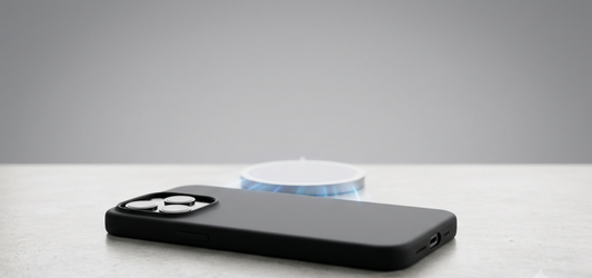 A wide product shot showing a black silicone case on a modern smartphone, angled on a polished concrete surface. Faint blue magnetic force lines visualize the connection attempt with a central white circular MagSafe charging pad.